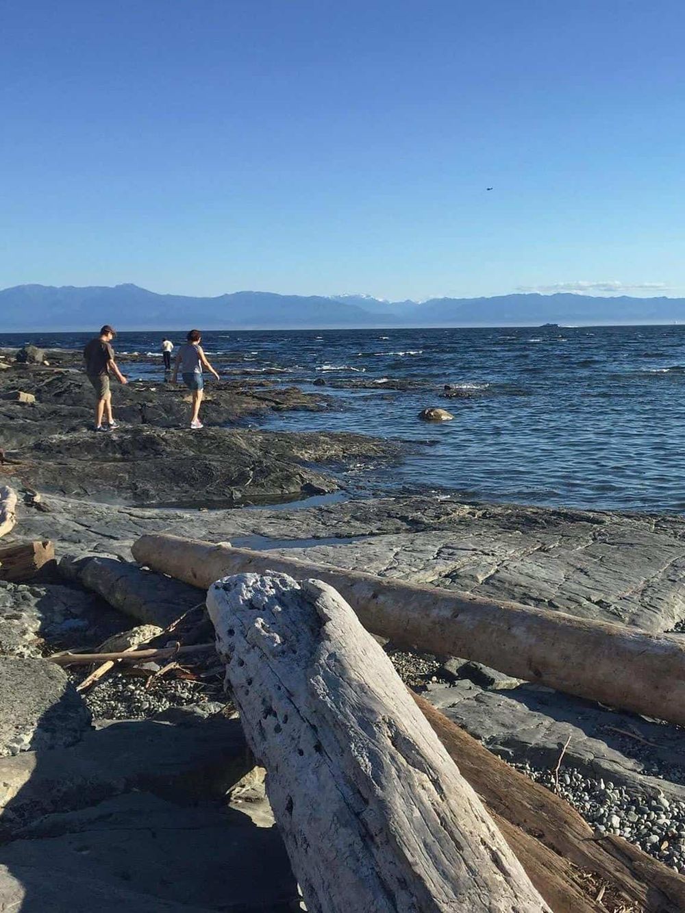 Scenic coastal view with people exploring the rocky shoreline and distant mountains under clear blue skies.