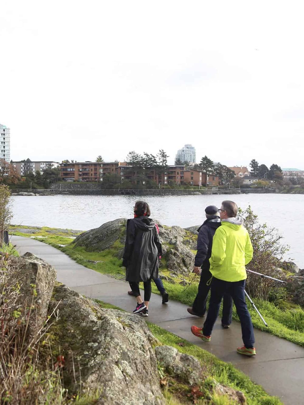 Serene waterfront park with people walking along the paved trail, overlooking city skyline and river.