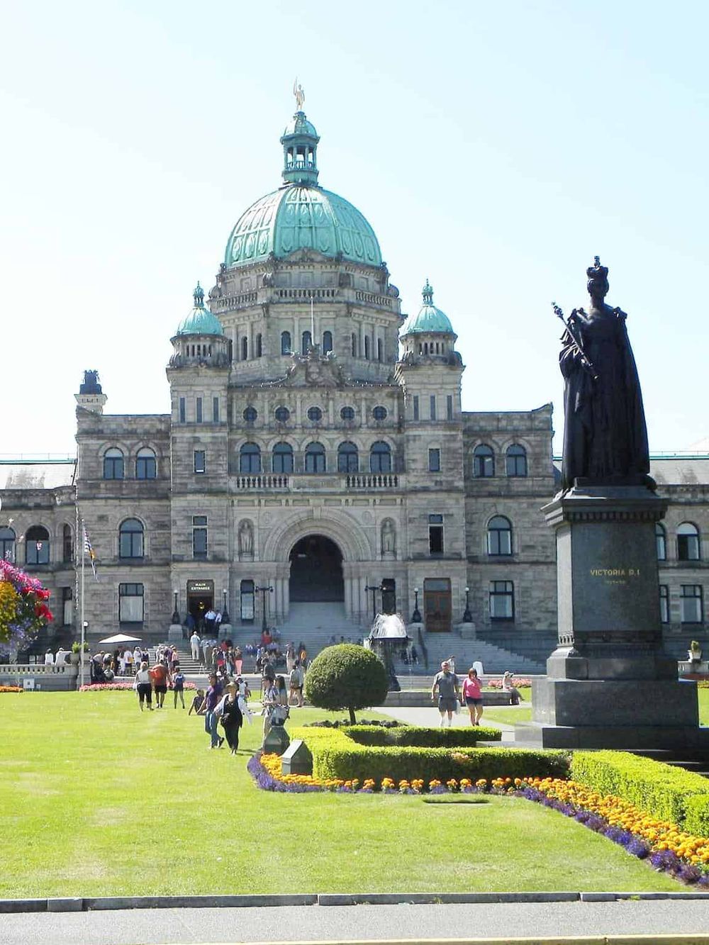 Historic government building with green domes in Victoria, Canada, featuring tourists and beautiful flower gardens.