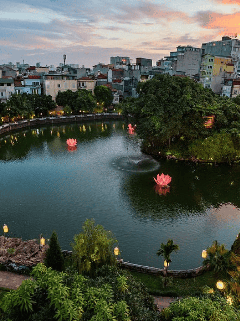 Serene pond with floating pink lotus flowers and cityscape at sunset in Hanoi, Vietnam.