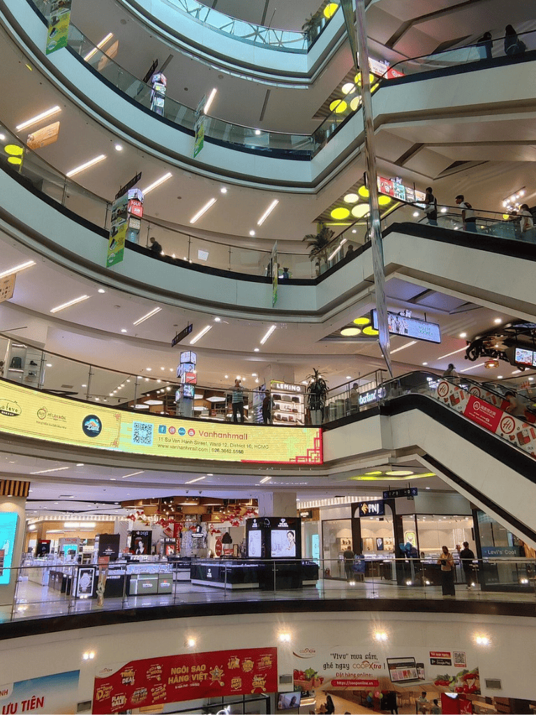Bright multi-level shopping mall interior with escalators, digital ads, and retail stores.