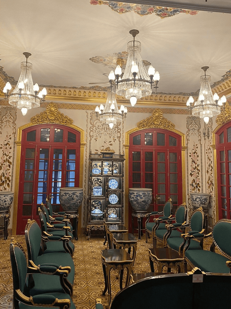 Lavish antique-style room with ornate chandeliers, green velvet chairs, and decorative porcelain display cabinets.