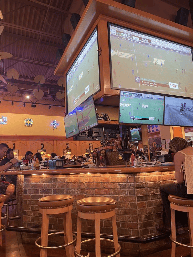 Upscale sports bar with multiple large screens showing live soccer matches, cozy brick counter, and guests enjoying drinks and food.