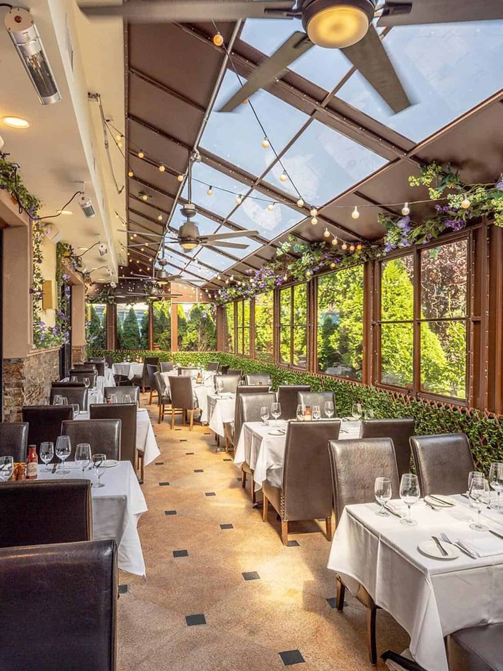 Bright indoor restaurant with glass roof, string lights, greenery, and elegant table settings for dining.