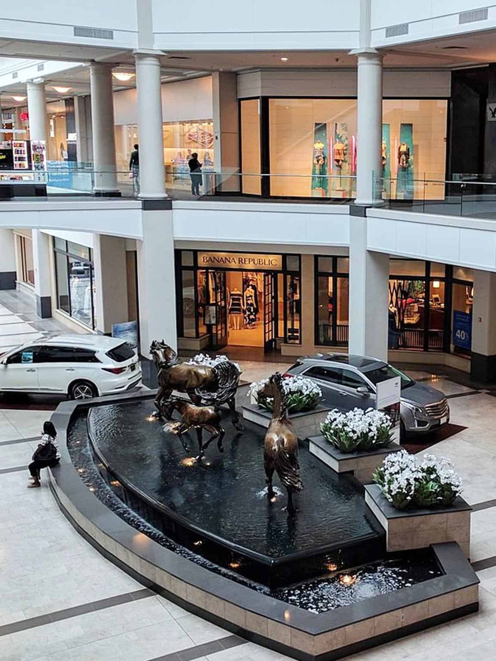 Luxurious shopping mall fountain with bronze horse sculptures and high-end retail stores.