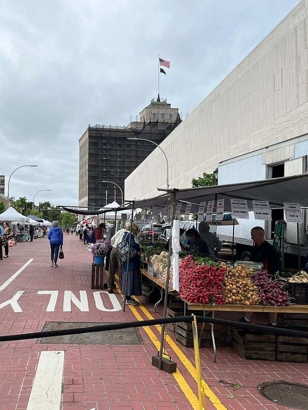 Fresh produce market with vendors selling vegetables and flowers in front of a city building, vibrant local market scene.