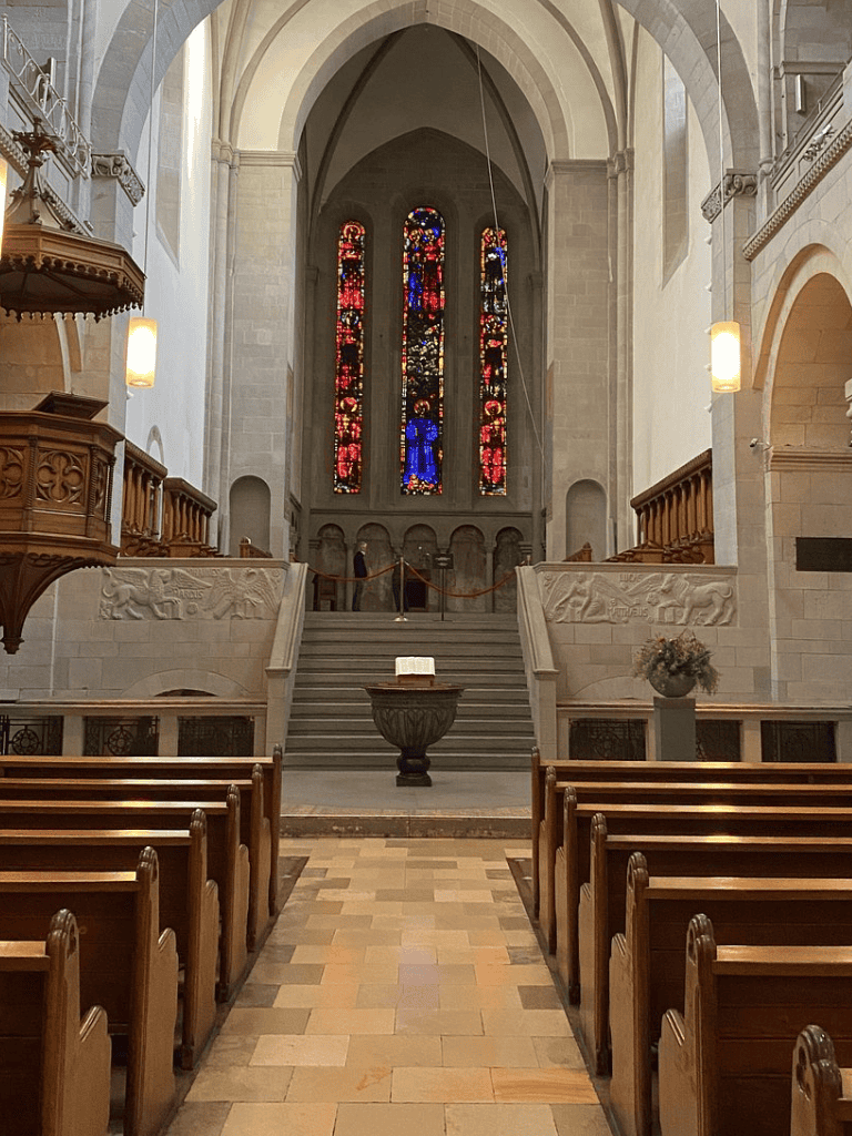 Stained glass window in a church interior with wooden pews and stone architecture.