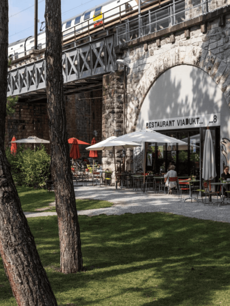 Outdoor restaurant with stone building, umbrellas, and train passing overhead in an urban setting.