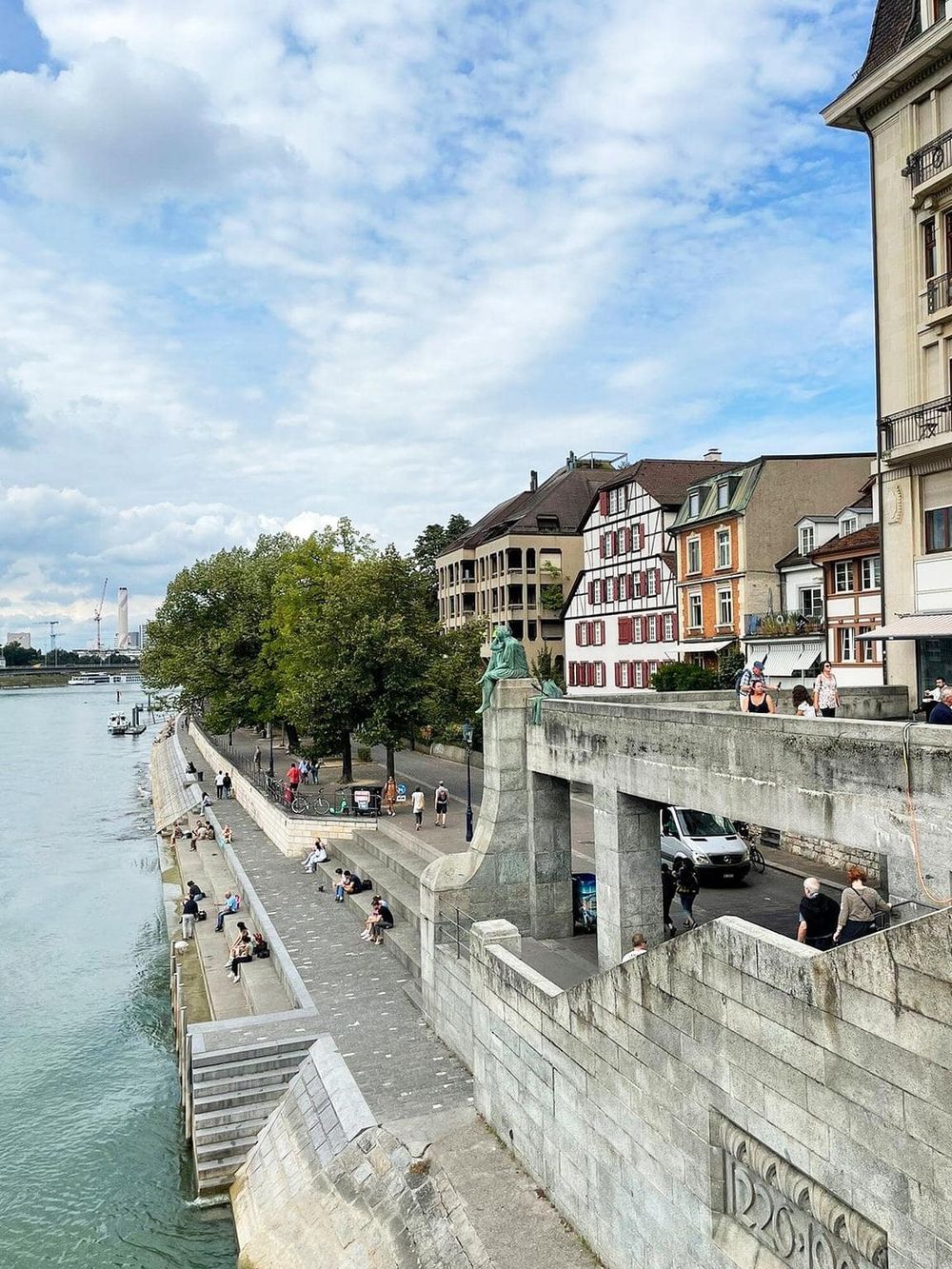 Scenic riverside walkway with historic buildings and people strolling near the water, capturing a vibrant city atmosphere.