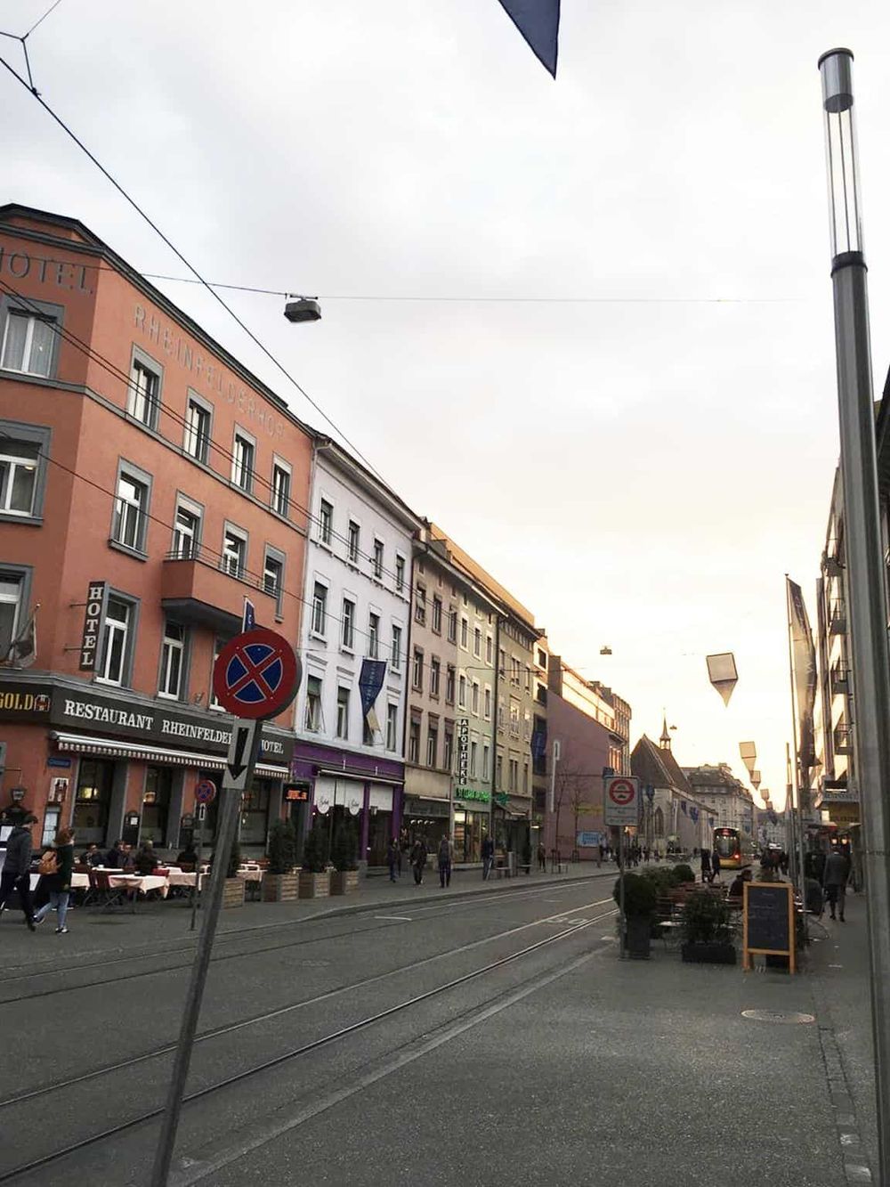 Colorful city street with hotels, restaurants, tram tracks, and pedestrians, in an urban European setting.