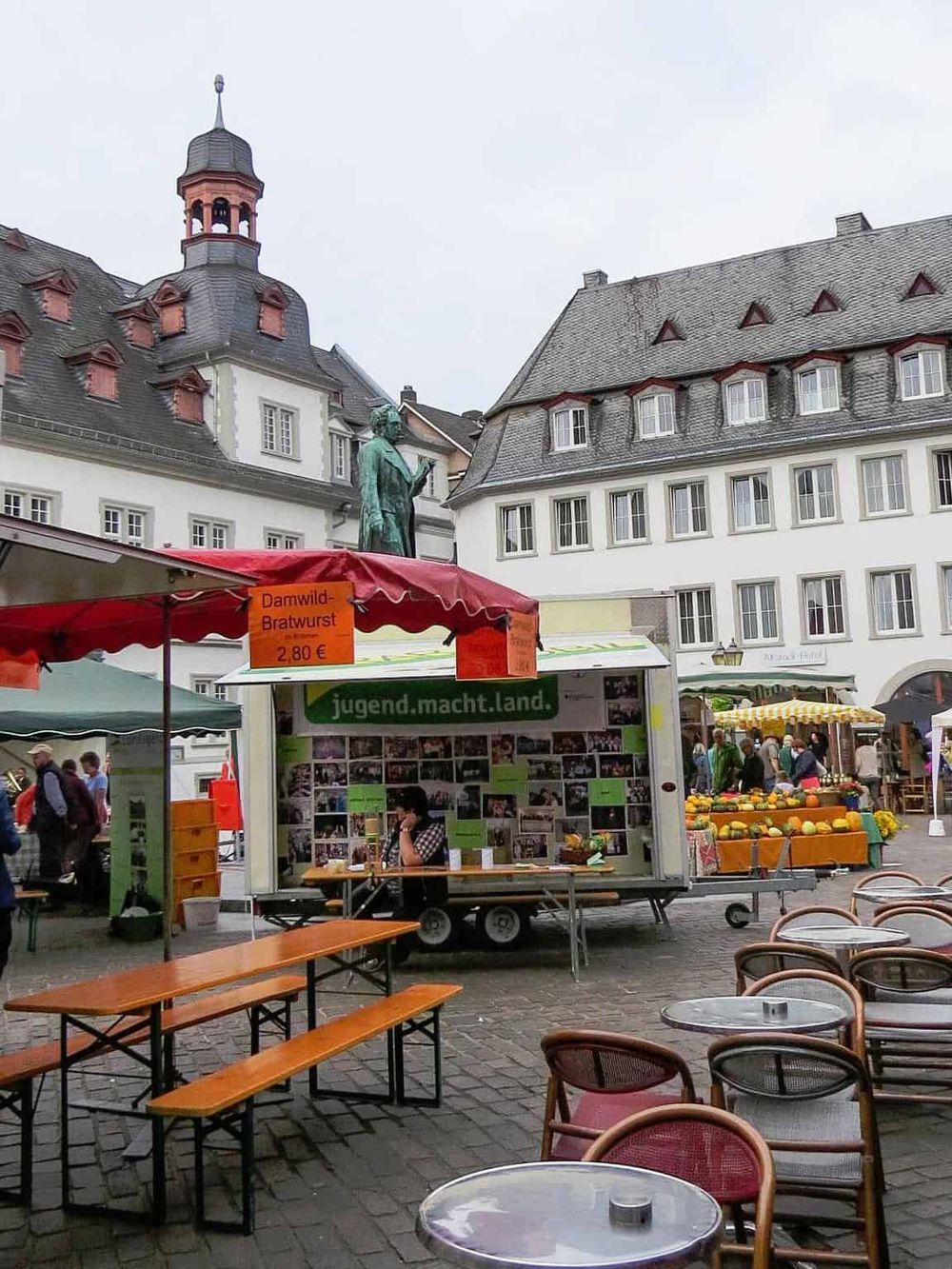 Exterior of a lively marketplace with food stalls, outdoor seating, historic European buildings, and a statue, capturing a vibrant local scene.