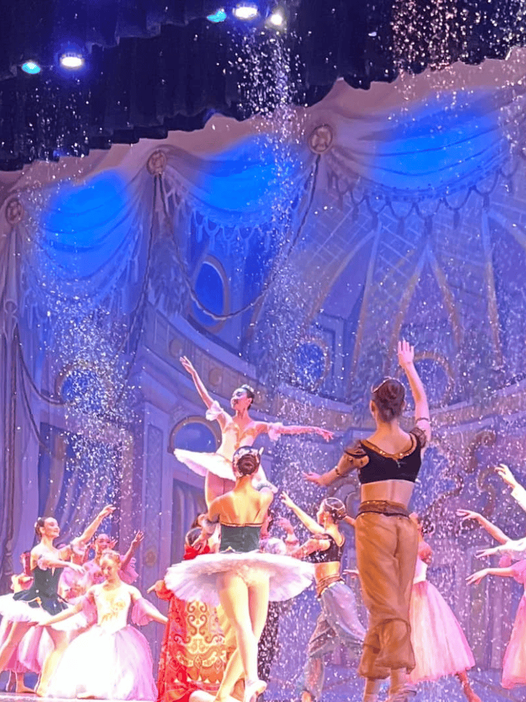 Elegant ballet dancers performing on stage with theatrical lighting and snow effects at Quest For Directions.