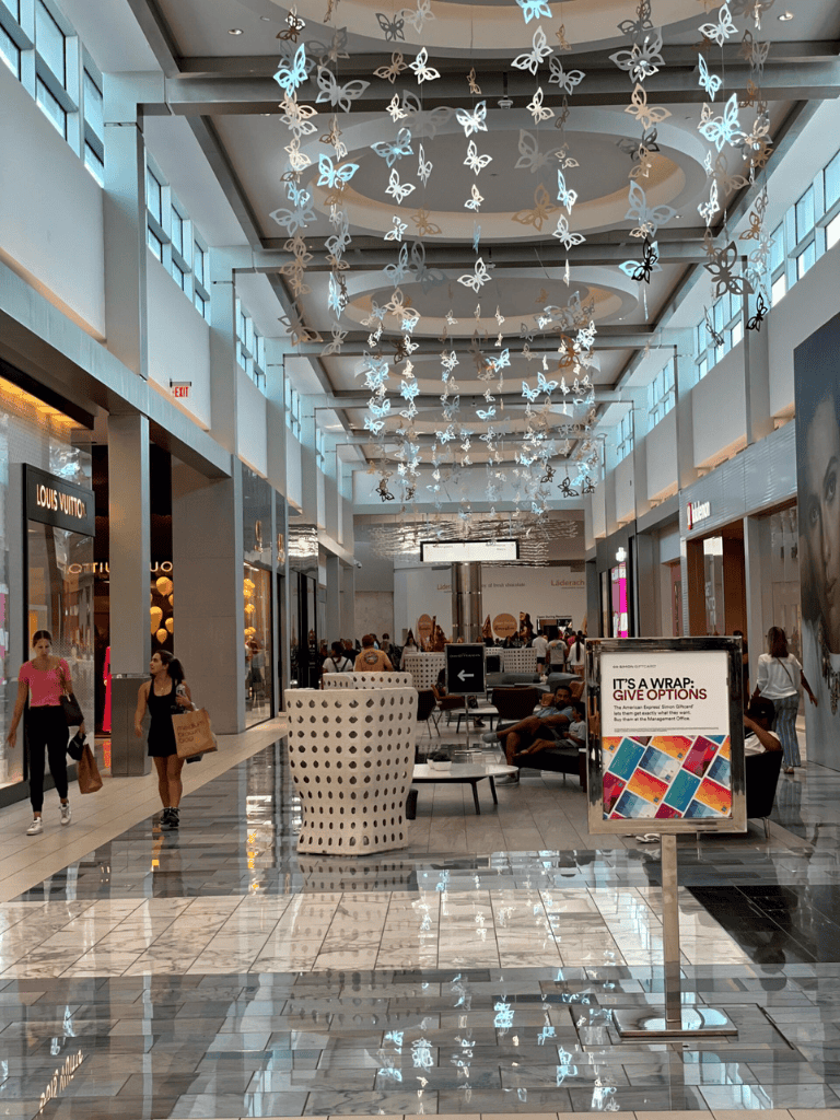 Shopping mall interior with butterfly ceiling decorations, shoppers, and retail stores.