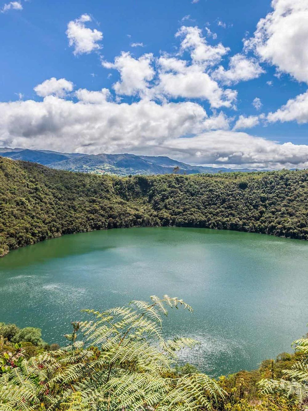 Serene lake surrounded by lush green hills and vibrant blue sky with fluffy clouds in the background.