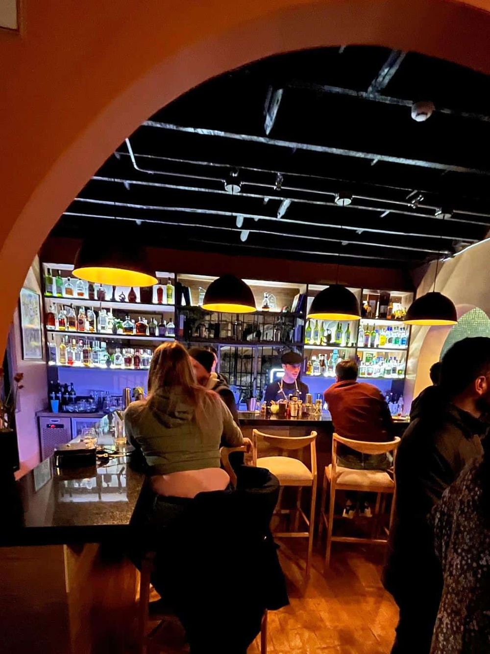 Cozy bar scene with customers enjoying drinks at a modern, dimly lit lounge; illuminated liquor shelves enhance inviting atmosphere.