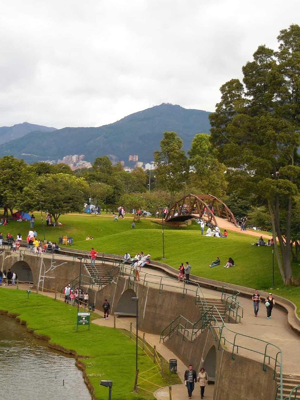 Bright city park with people relaxing, walking, and enjoying nature near a river and mountain backdrop.