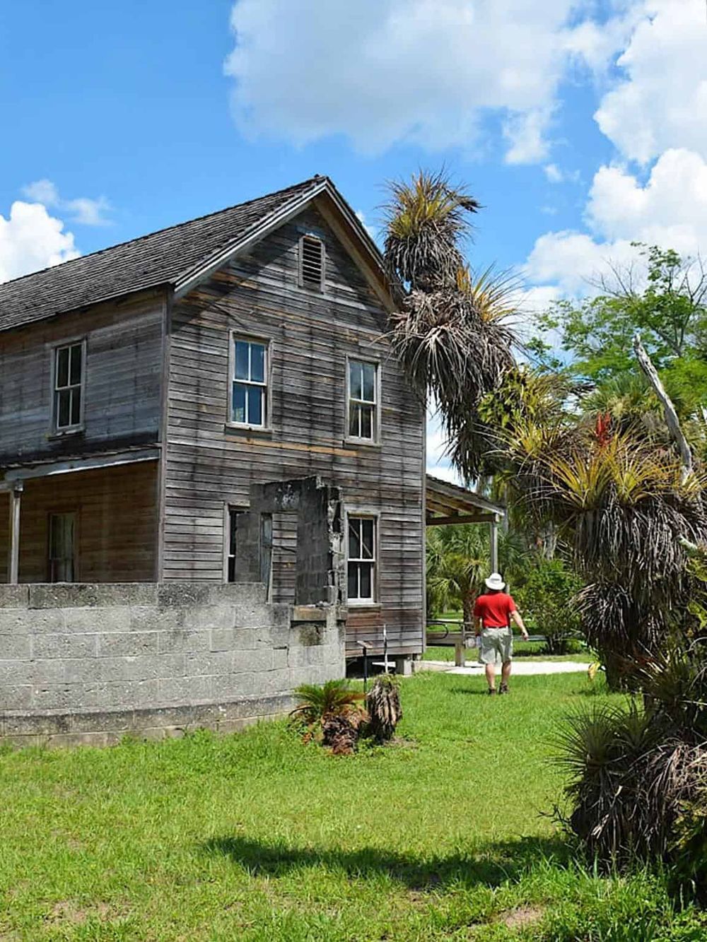 Old wooden house with tropical trees, sunny sky, and a person walking away, representing historic buildings and travel destinations.