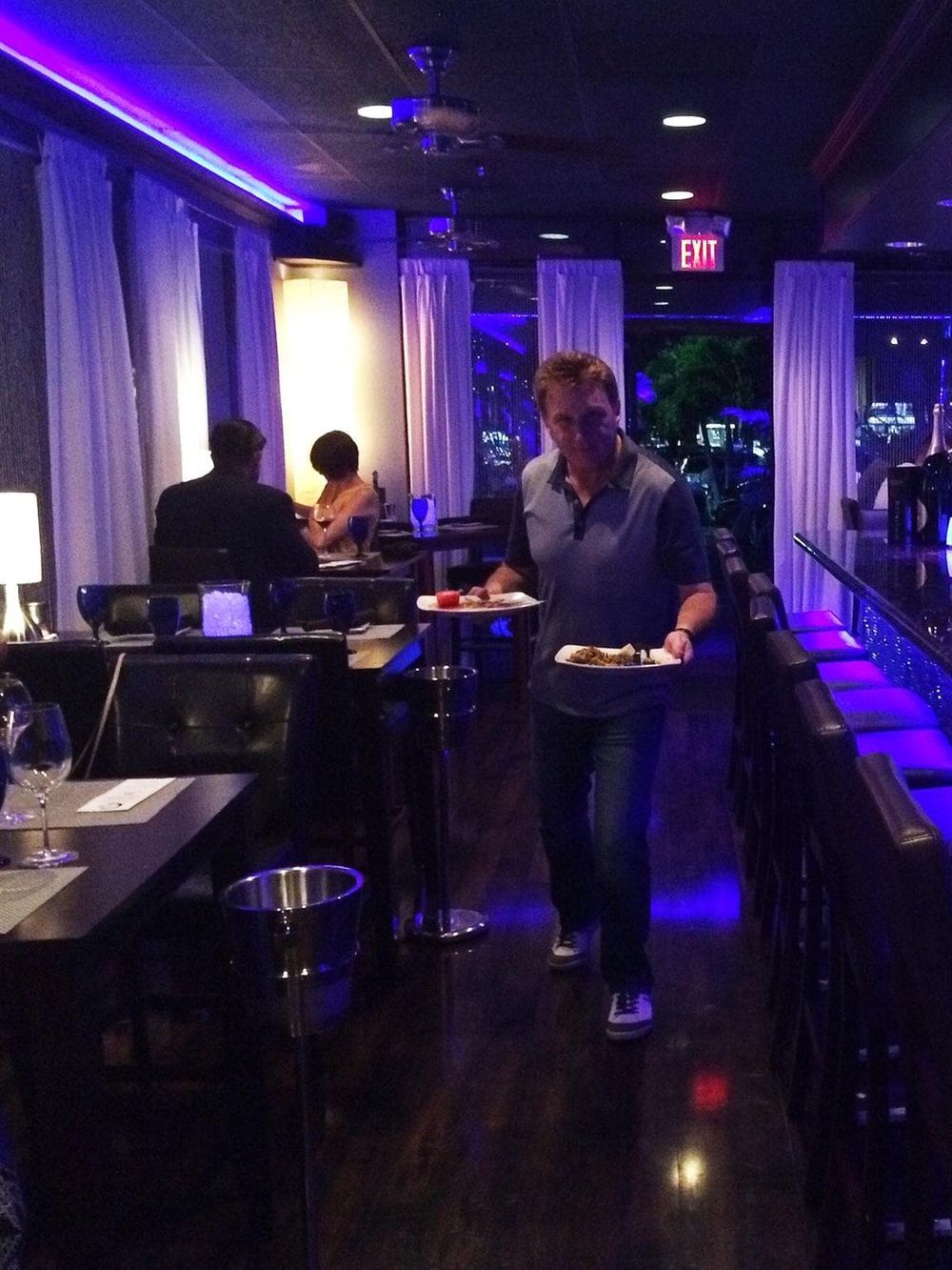 Elegant restaurant scene with guests dining and a server carrying plates, illuminated by ambient blue lighting.