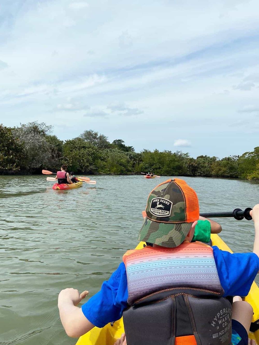 Kayaking adventure on scenic waterway with family, surrounded by lush green trees and peaceful blue sky.
