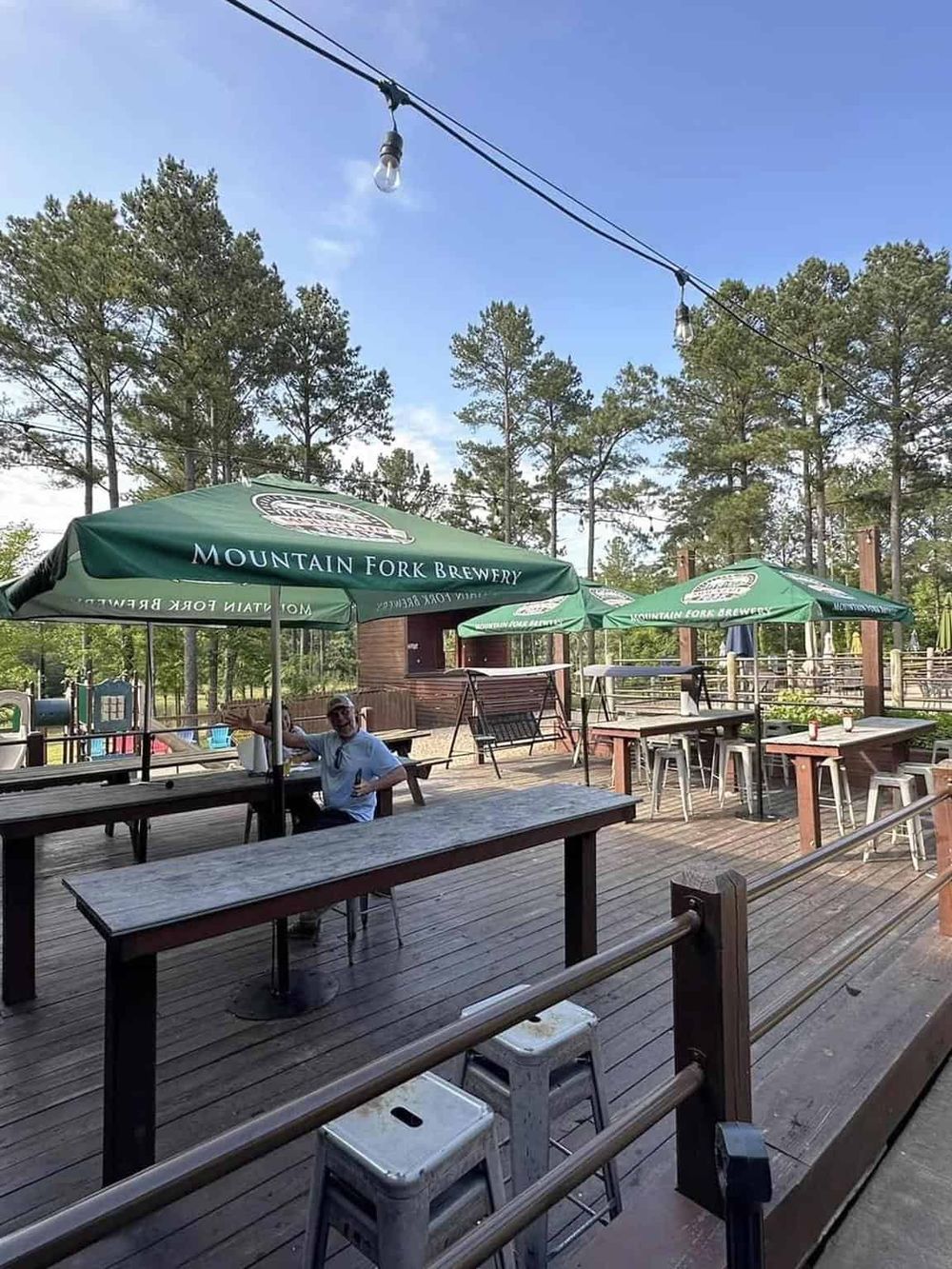 Outdoor patio with Mountain Fork Brewery umbrellas and seating, surrounded by tall pine trees.