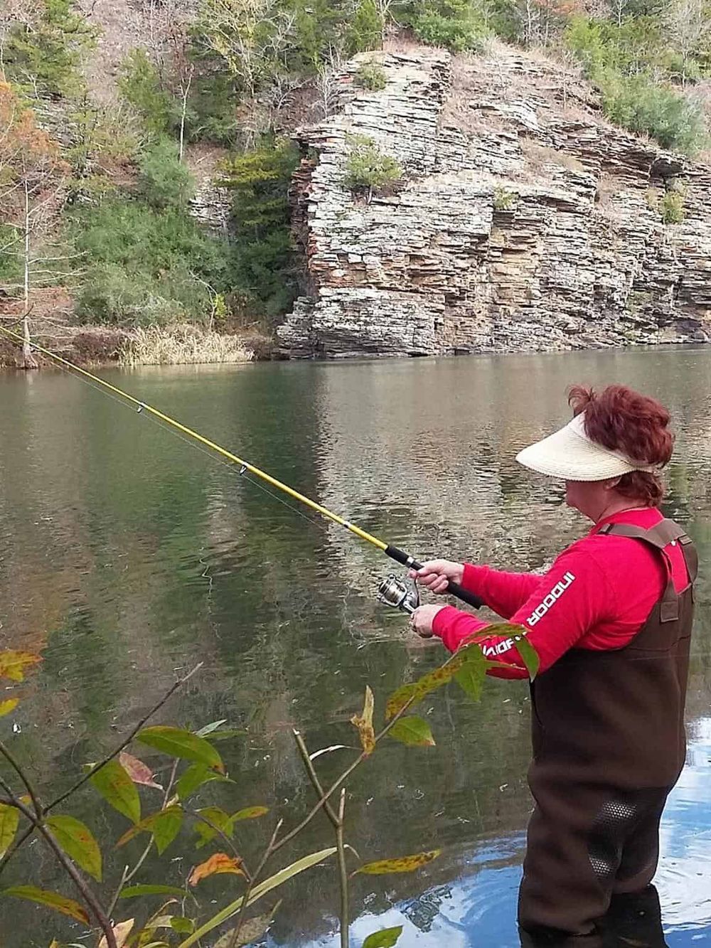 Trout fishing woman with hat in freshwater lake surrounded by trees and rocky cliffs.