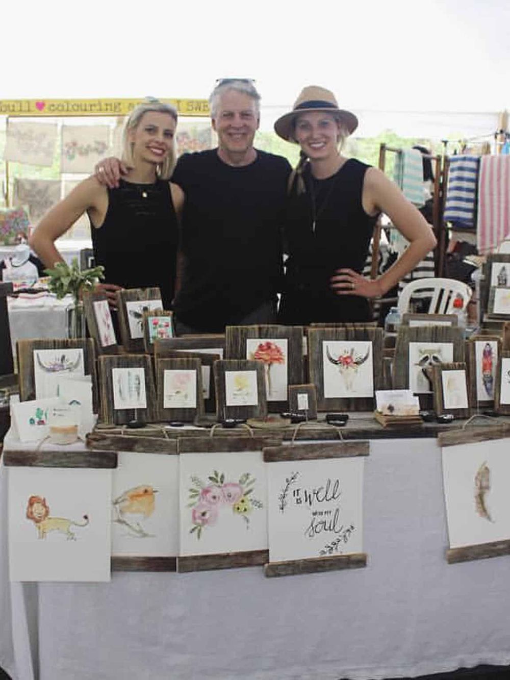 Colorful art booth at craft fair with smiling vendors, showcasing succulent watercolor paintings for sale.
