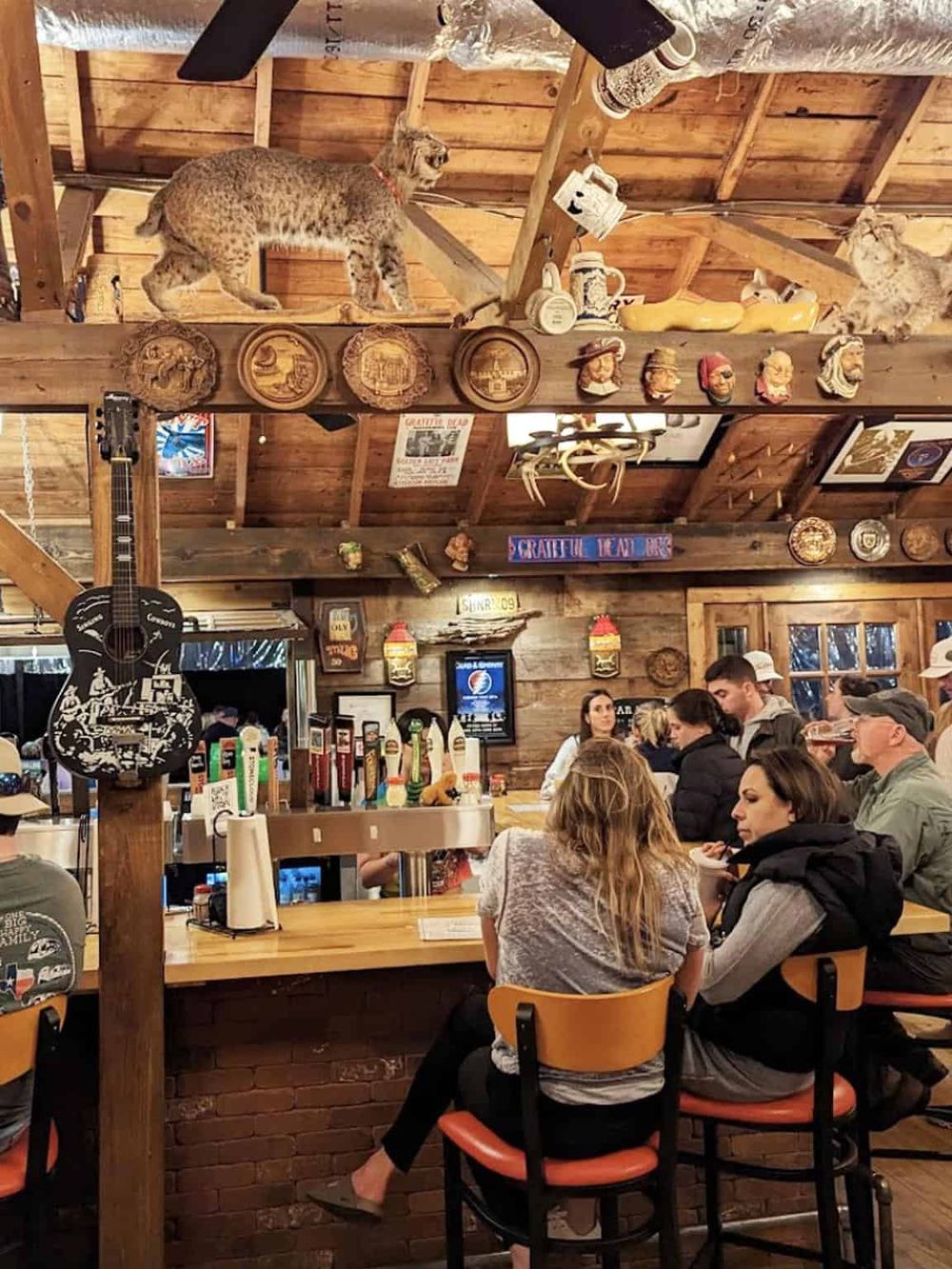 Cozy rustic bar with wood decor, mounted animal trophies, and celebrity masks, filled with patrons enjoying drinks and conversation.