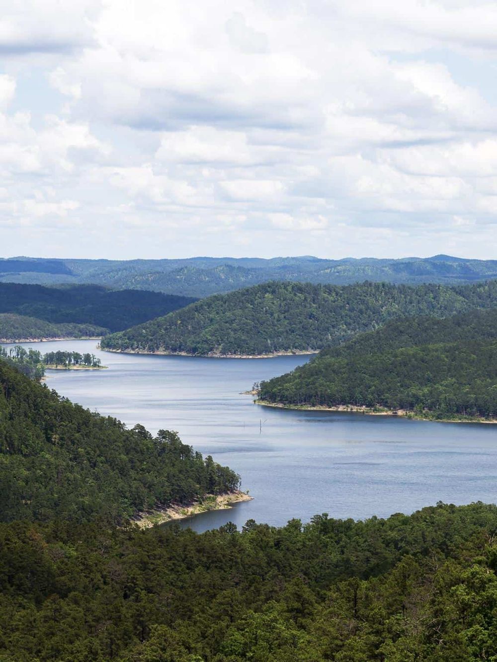 Vast reservoir surrounded by lush green hills and forested landscape under cloudy sky.