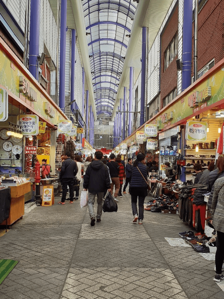 Vibrant indoor shopping market with diverse vendors and shoppers, featuring a glass arched ceiling for natural light.