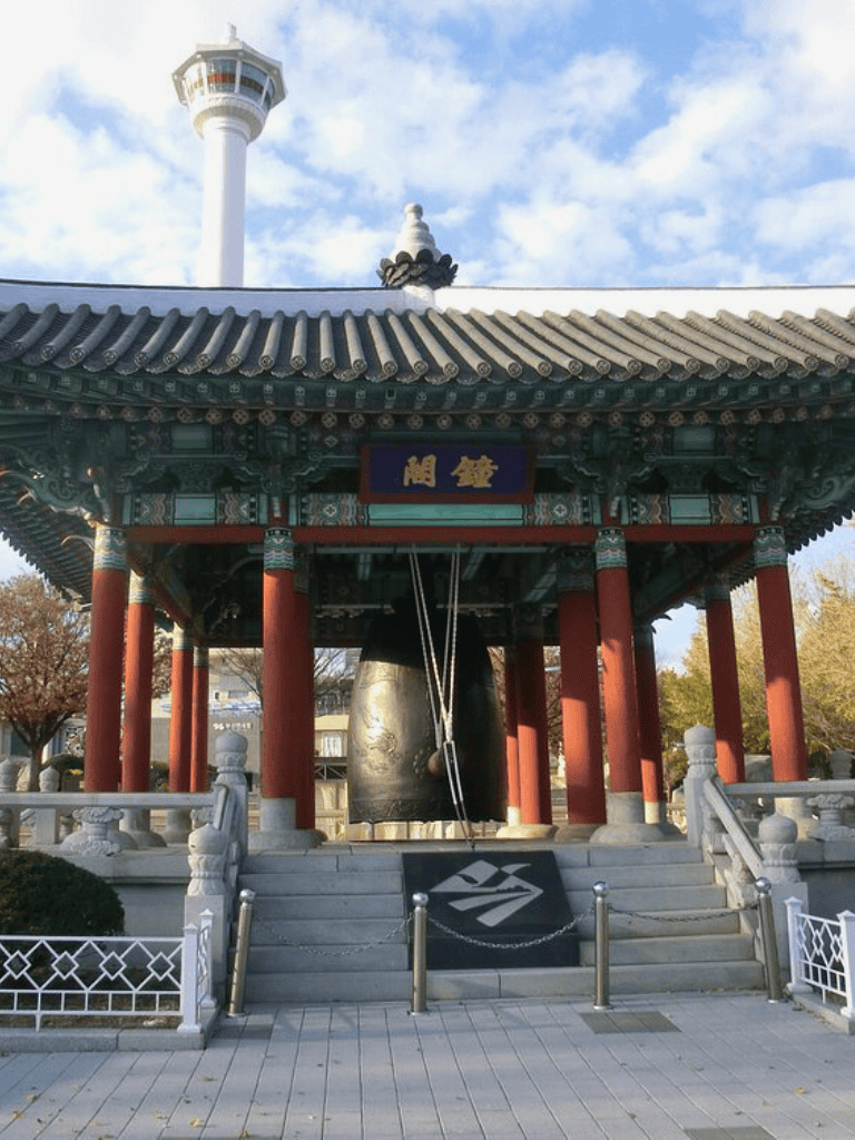 Traditional Korean bell pavilion with a large hanging bell and modern observation tower in background.