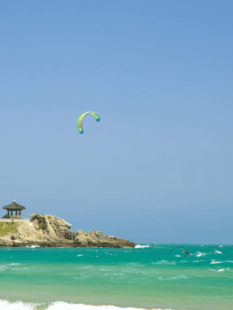 Colorful kite flying over ocean waves near rocky coastline with traditional pavilion.