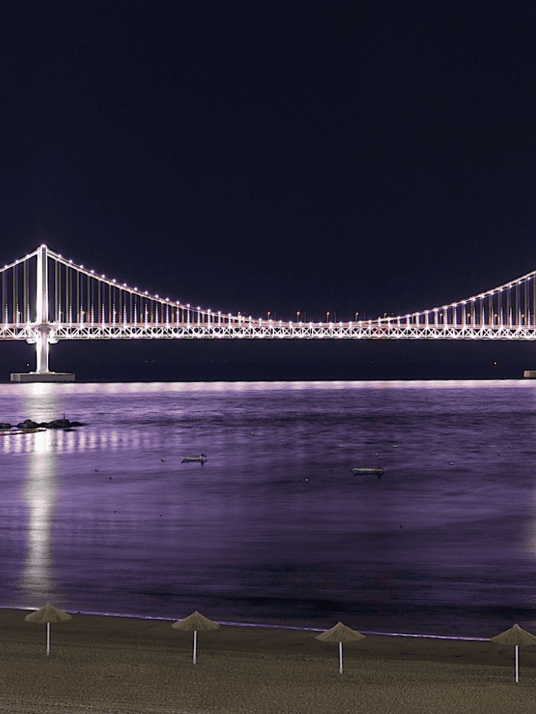 Brightly lit suspension bridge at night over calm water, with a sandy beach and umbrellas in the foreground.