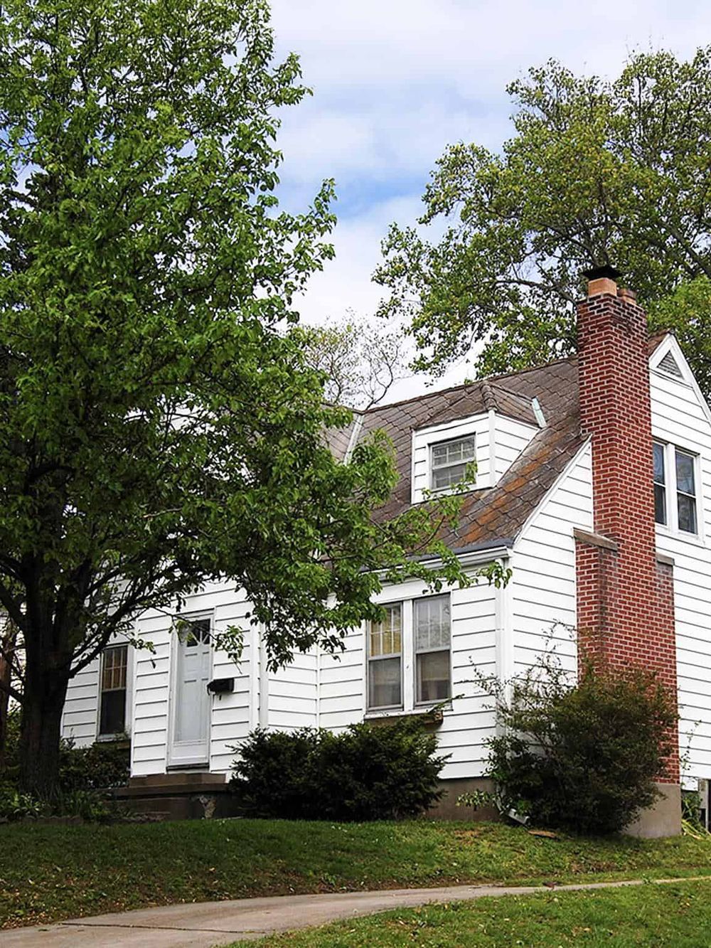 Charming white house with red brick chimney, surrounded by lush green trees and bushes, in a scenic suburban neighborhood.