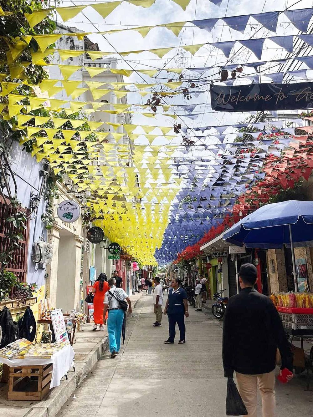 Colorful decorative street with yellow, blue, and red banners, lively shops, and pedestrians in a vibrant urban setting.