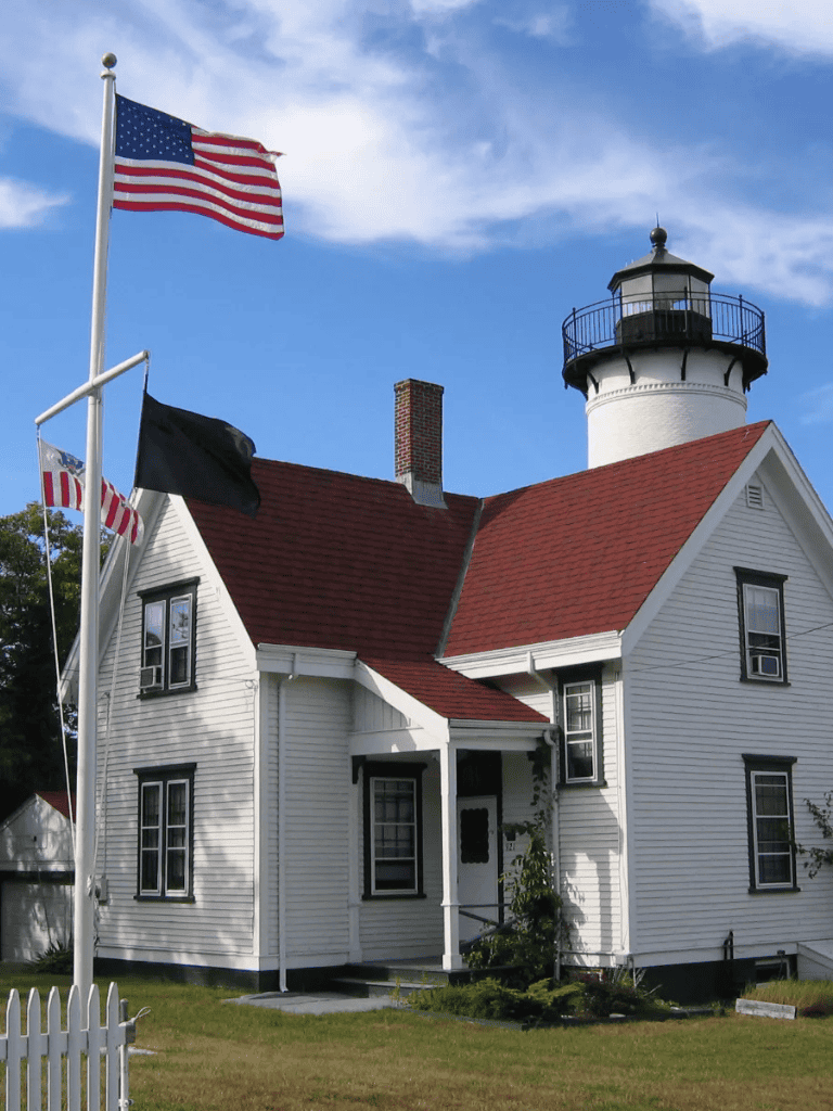 American flag flying in front of a classic lighthouse and historic house, coastal navigation and maritime history.