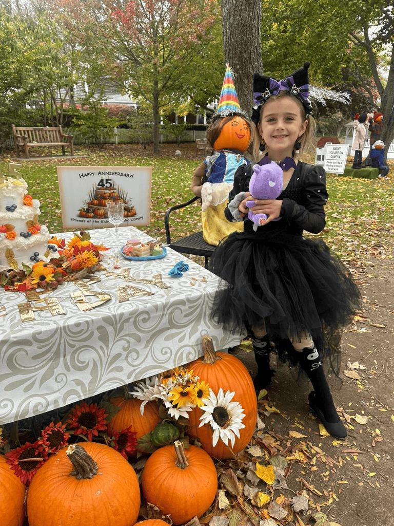 Festive girl in Halloween costume holding plush toy at pumpkin themed event decorated with gourds and fall flowers.