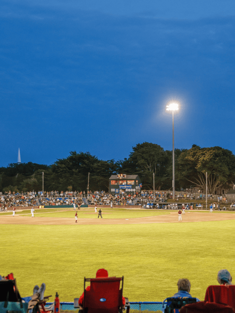 Brightly lit baseball stadium during night game with spectators and players on the field.