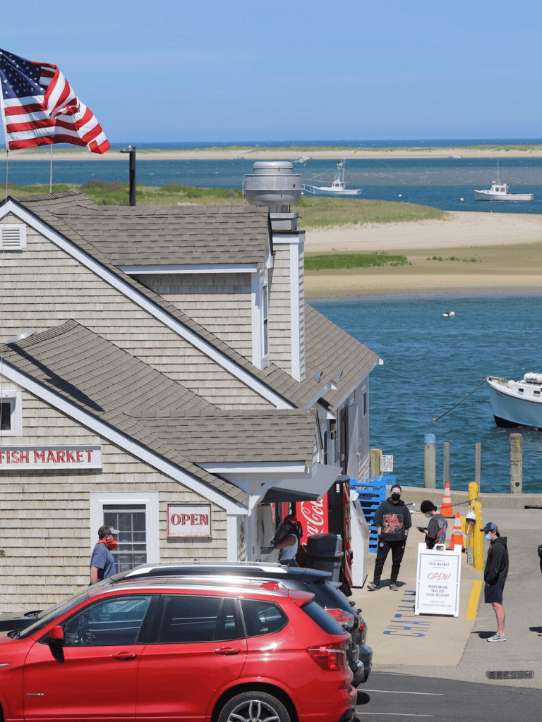 American flag flying over coastal waterfront with boats, harbor, and seaside buildings.