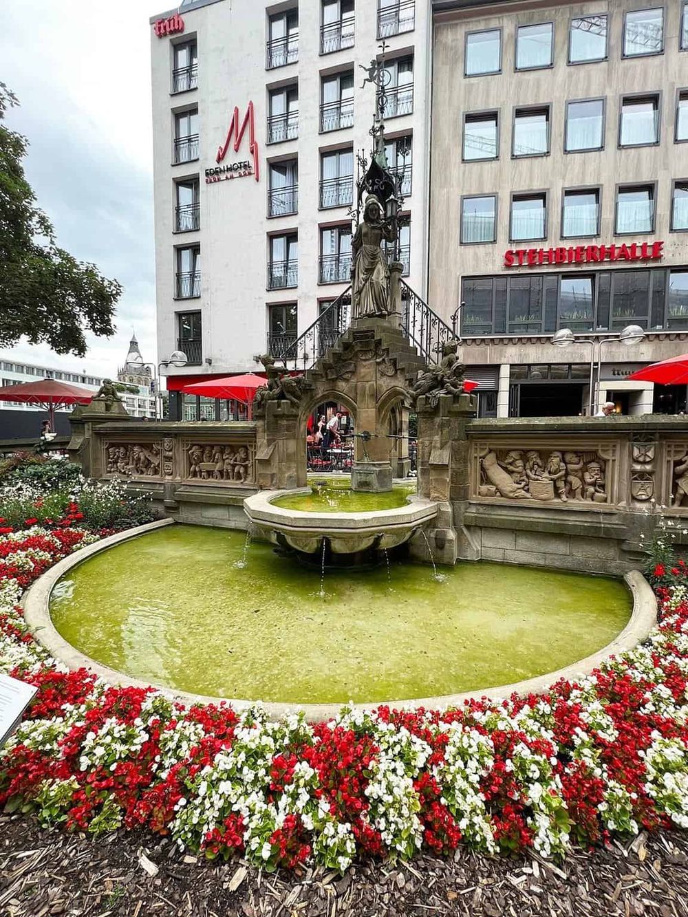 Beautiful historic fountain with intricate sculptures in an urban setting, surrounded by colorful flowers and modern buildings.