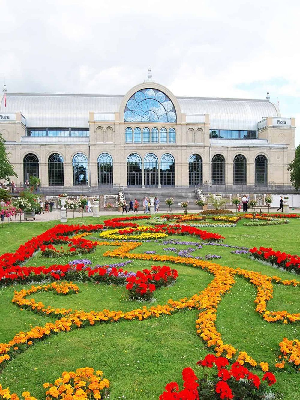 Colorful flower garden in front of historic conservatory building with large glass windows.