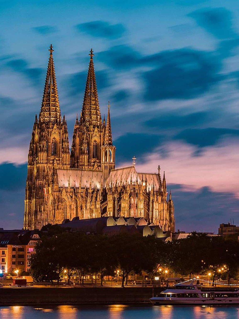 Gothic cathedral illuminated at dusk with river and city skyline in the foreground.