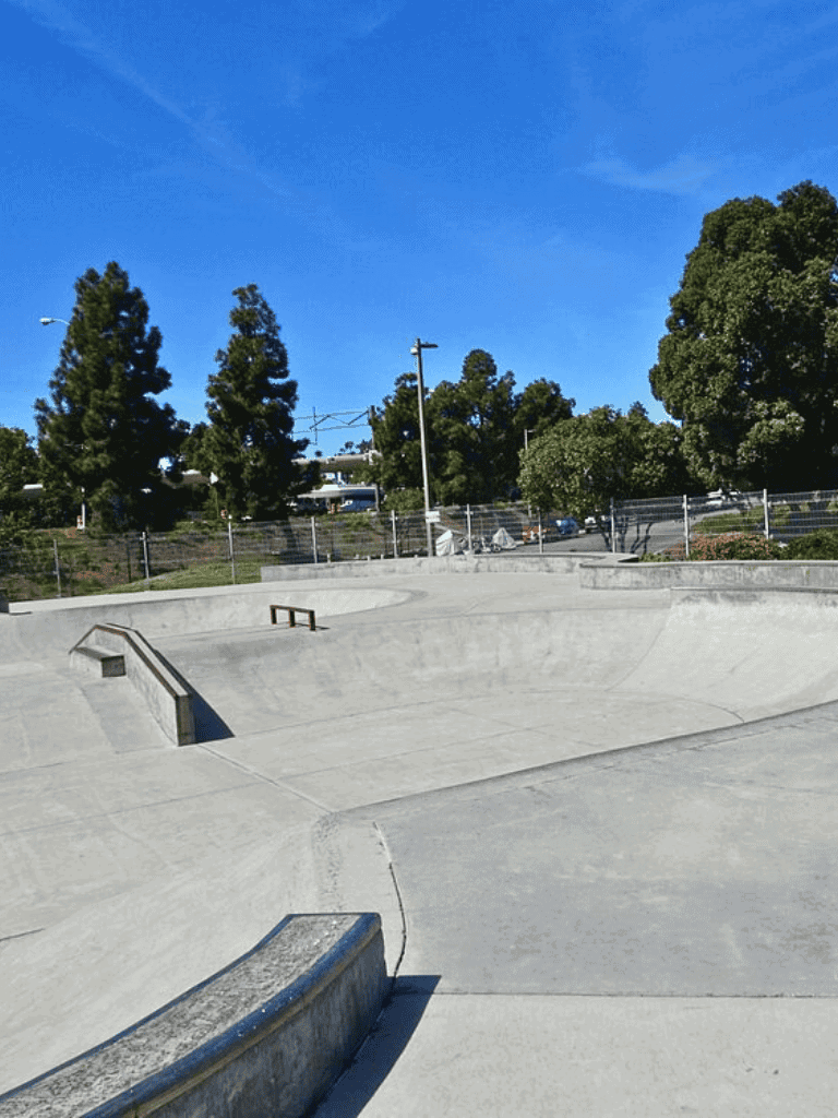 Smooth concrete skateboard park with ramps and rails, sunny day, green trees in the background.