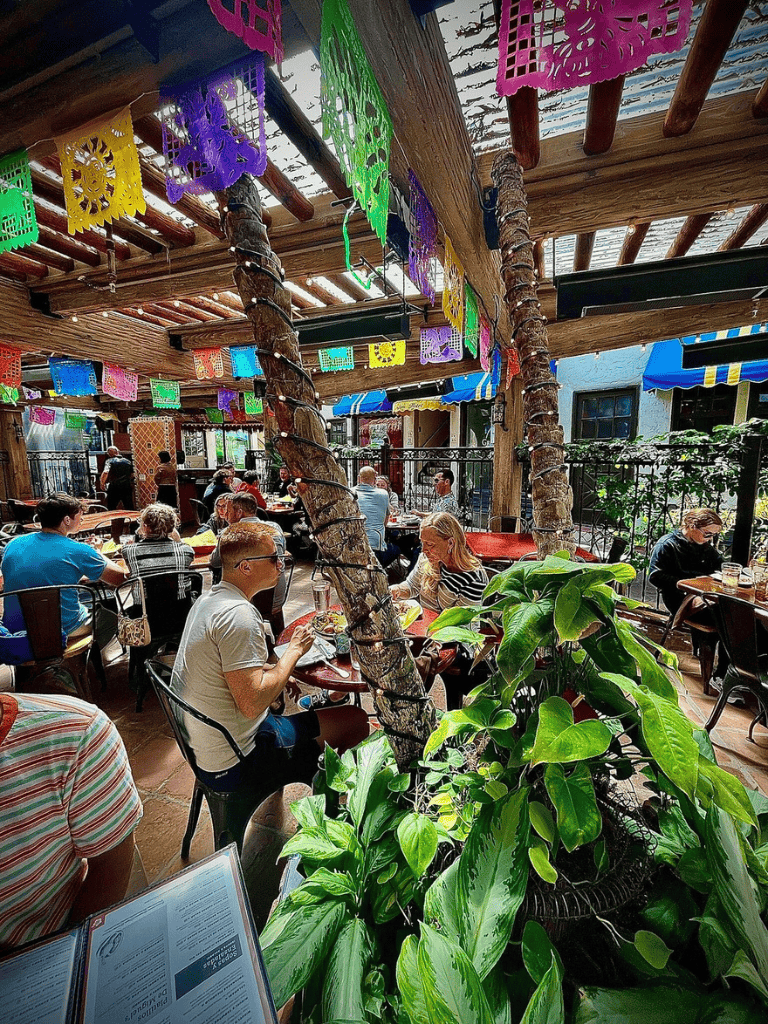 Colorful papel picado decorations inside a vibrant, lively restaurant with outdoor seating and lush greenery.
