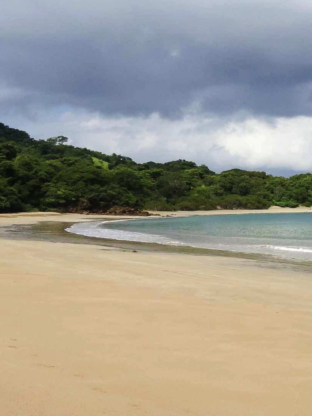 Serene beach scene with lush green hills, sandy shore, and cloudy sky in the background.