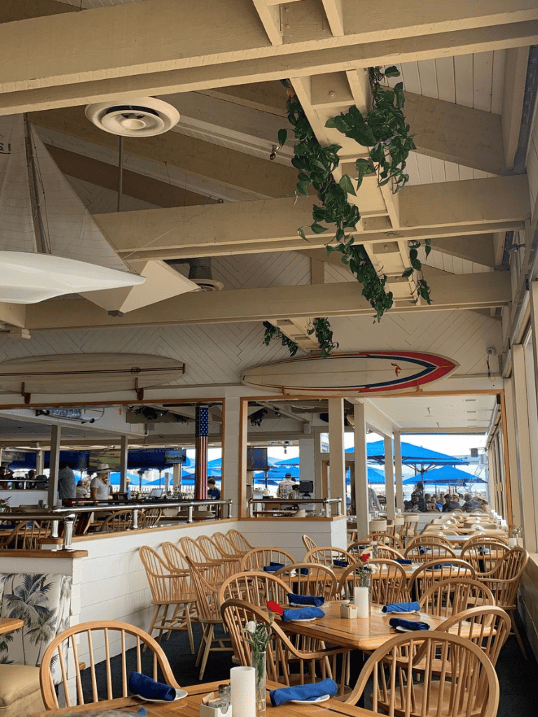 Bright seaside restaurant with wooden tables, blue napkins, surfboards, and outdoor ocean views.