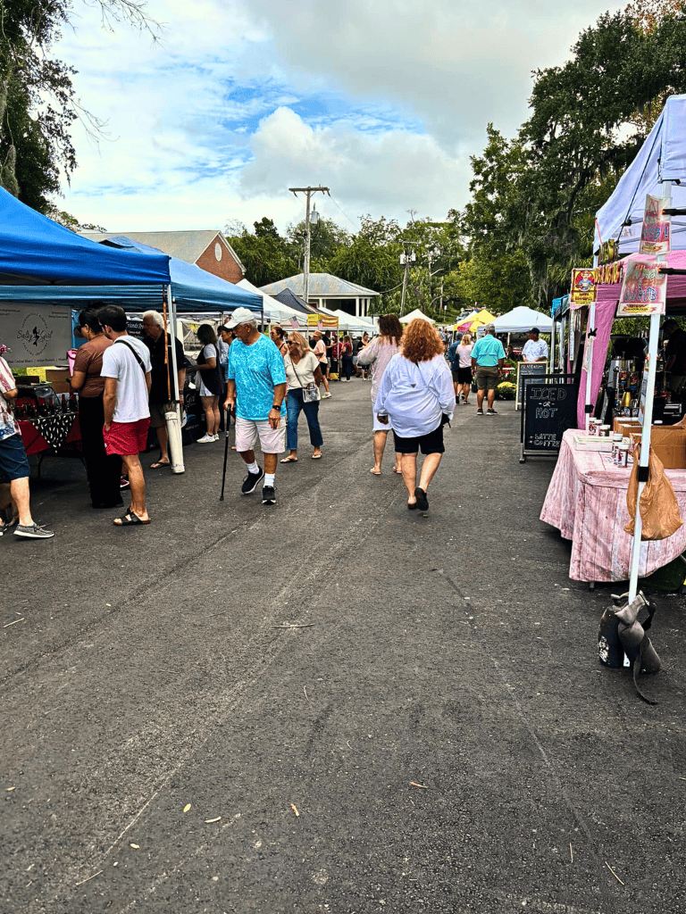 Colorful outdoor market with diverse vendors and shoppers under blue tents, lively atmosphere, sunny sky, and lush trees.
