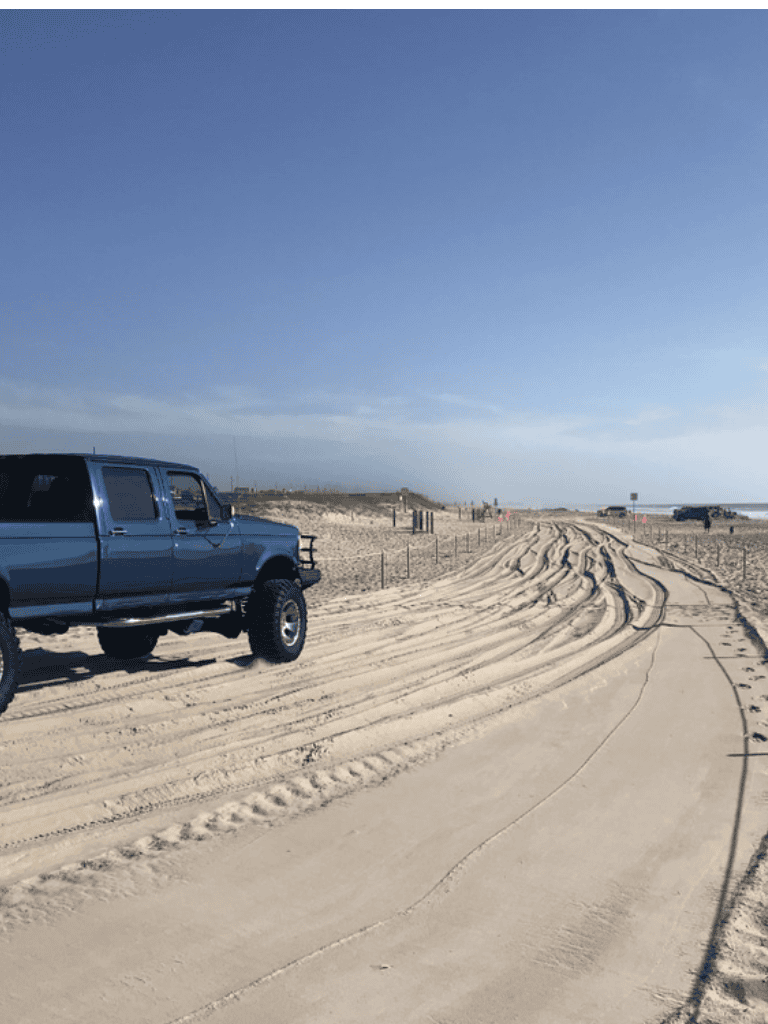 Off-road vehicle parked on beach with sandy dunes and tire tracks in the background.