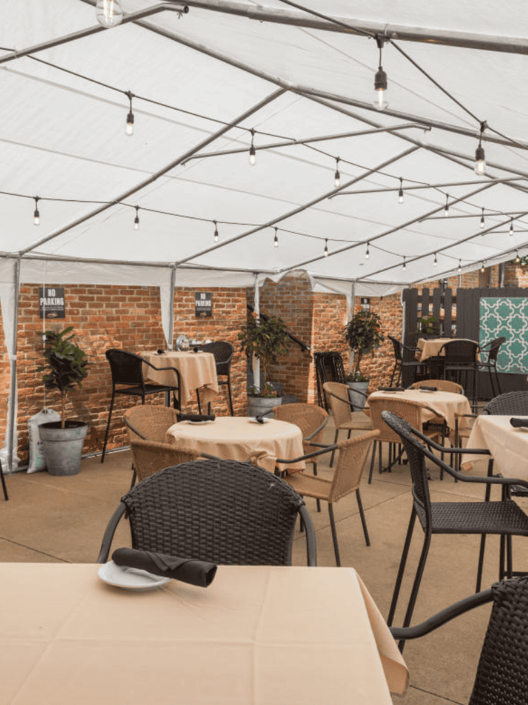 Elegant outdoor dining setup under a white tent with string lights and brick wall decor.