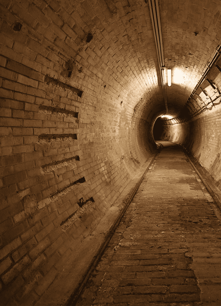 Underground brick tunnel with lighting and pipes, leading into darkness.