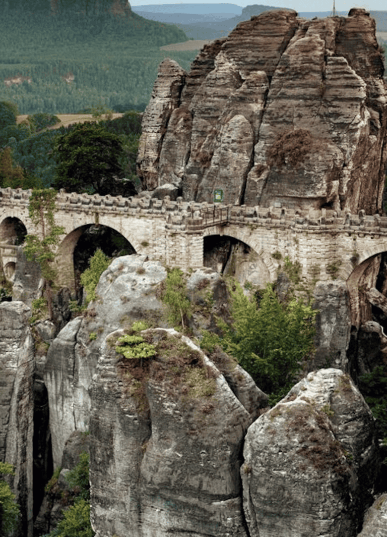 Ancient stone bridge over rocky canyon with lush green landscape in background.